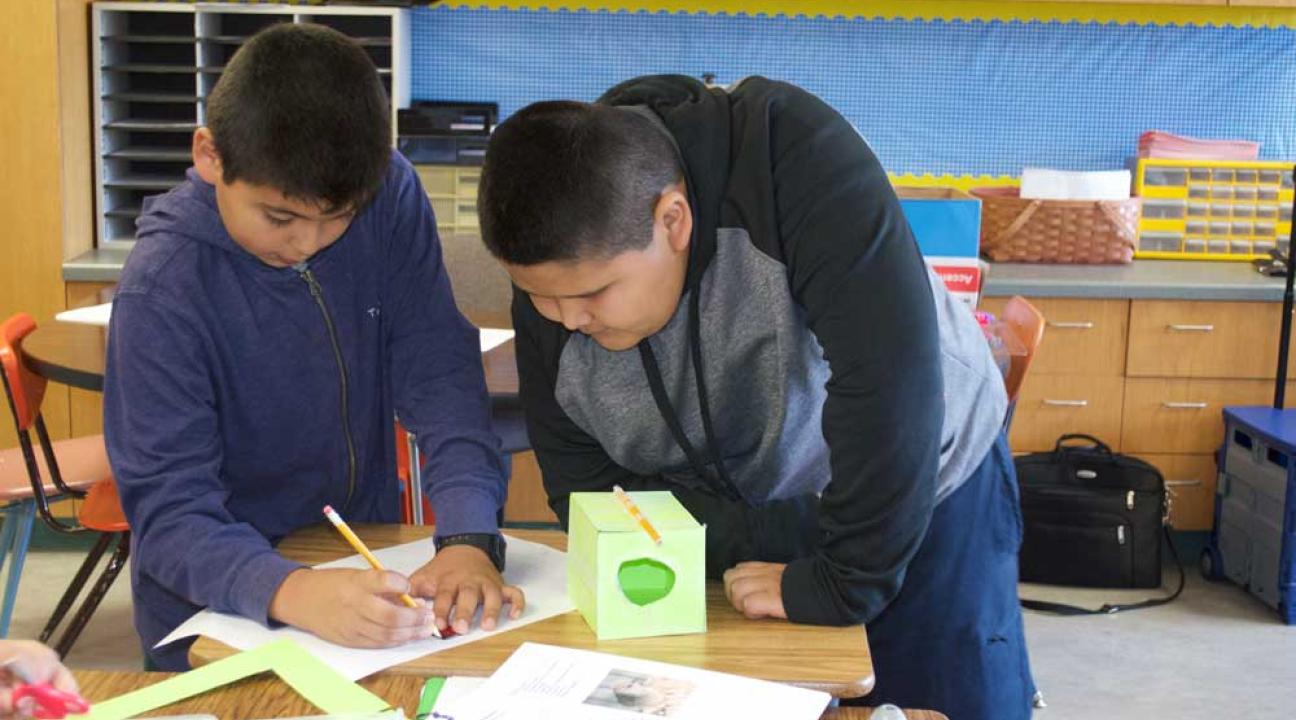 Two boys are hunched over a desk, one is writing notes, the other boy watches.