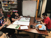 3 students site at a table.  One is using a laptop, one is writing, the third student is holding an electronic device.
