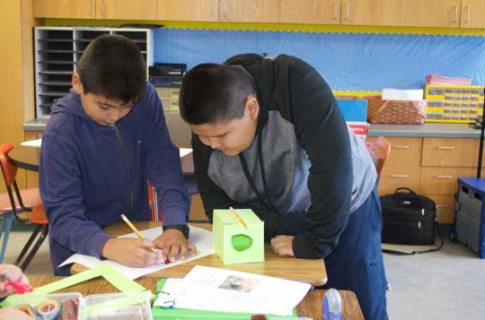 Two boys are hunched over a desk, one is writing notes, the other boy watches.