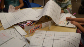 Graph paper on on a table.  Hands are writing on the graph.